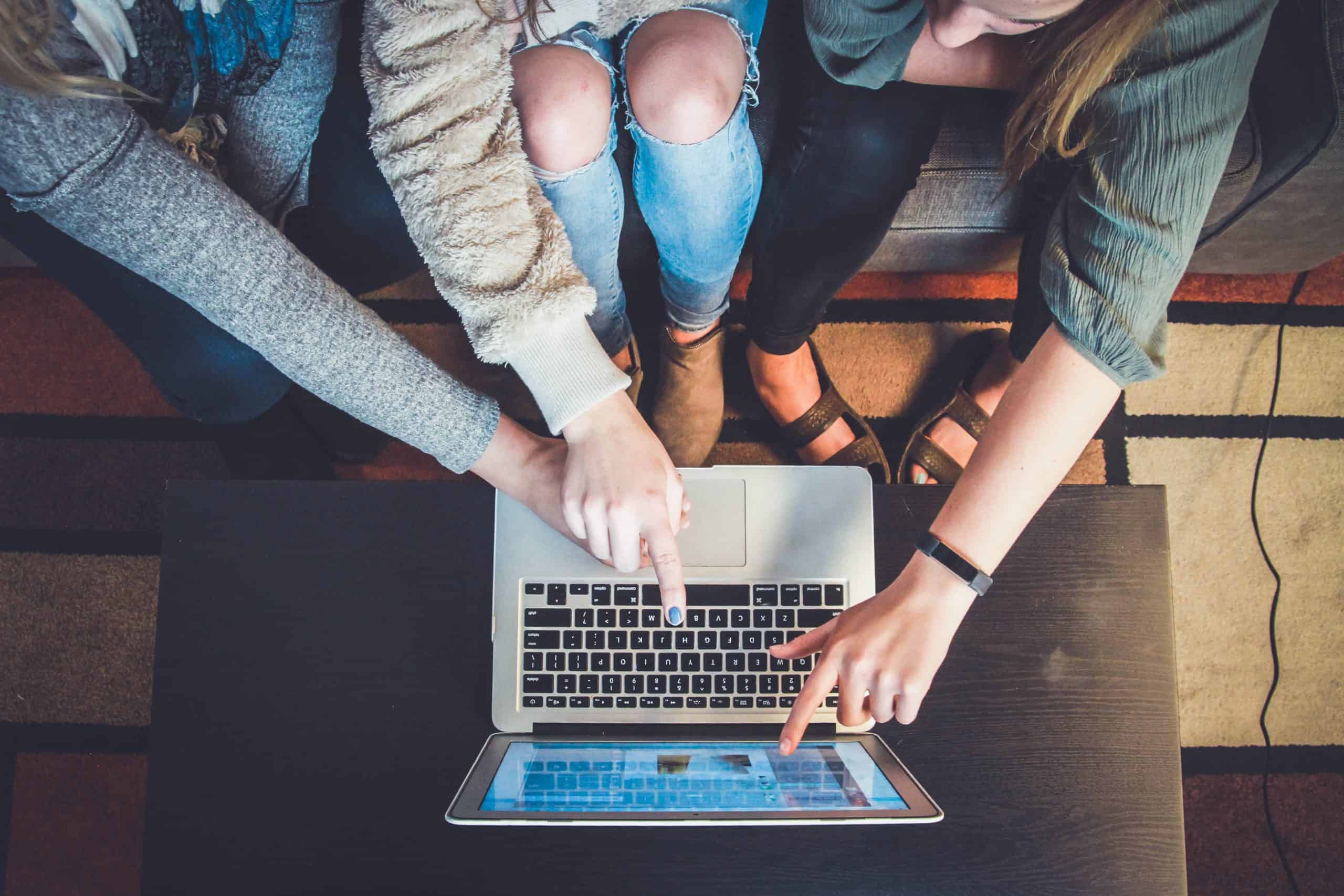This image is shot overhead and shows three women sitting on a couch in front of a laptop. The laptop is on the coffee table in front of them, and, although we can't see what's on it, the screen is lit up. Each girl is pointing at a different place on the screen, implying that they each have differing opinions/interests about what is on it.