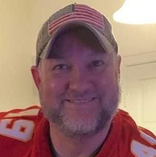 man in a red jersey wearing a beige hat with an American flag smiling to the camera
