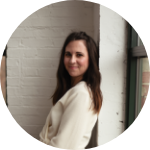 woman with brown hair wearing a white sweater and leaning on a wall smiling to the camera