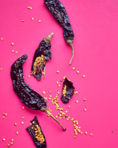 three dried chili peppers and seeds on a pink background