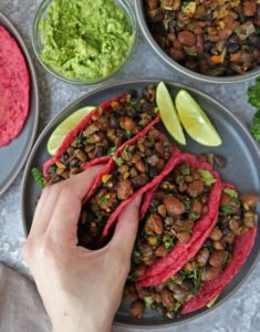 person grabbing a beet tortilla taco with bean filling from a plate of 4 tacos with sliced limed and a bowl of guacamole