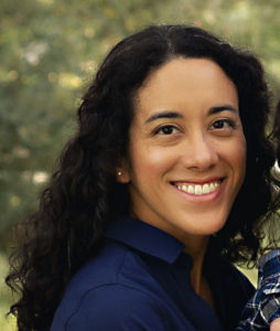 woman in a blue shirt with curly hair smiling at the camera with foliage behind her