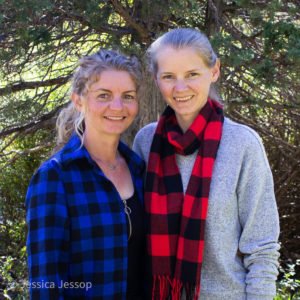 two women with blonde hair smiling in front of a tree. left woman on the left is wearing a blue paid button up and women on right wearing a gray sweater and red plaid scarf.