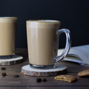 a vanilla latte in a clear coffee mug on a coaster with coffee beans and cookies on the brown table