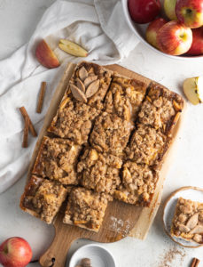 Slices of salted caramel apple pie bars on a charcuterie board surrounded by apples and cinnamon sticks