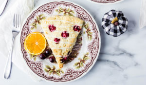 cranberry scone with white drizzle on a plate with a orange slice and cranberries next to a black and white pumpkin