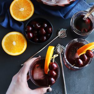 two red drinks with slices of oranges and cherries on a stick. half cut oranges and a bowl and cup of cherries on the side