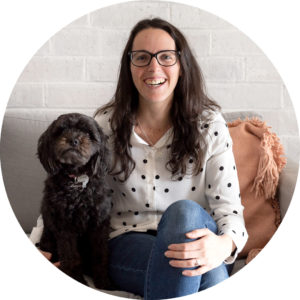 woman with brown hair, glasses, jeans, and a white polkadot shirt smiling with a dog