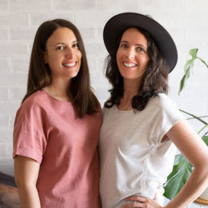 two women with brown hair smiling in front of a plant. left woman wearing a pink shirt and right woman wearing a gray shirt and brown hat