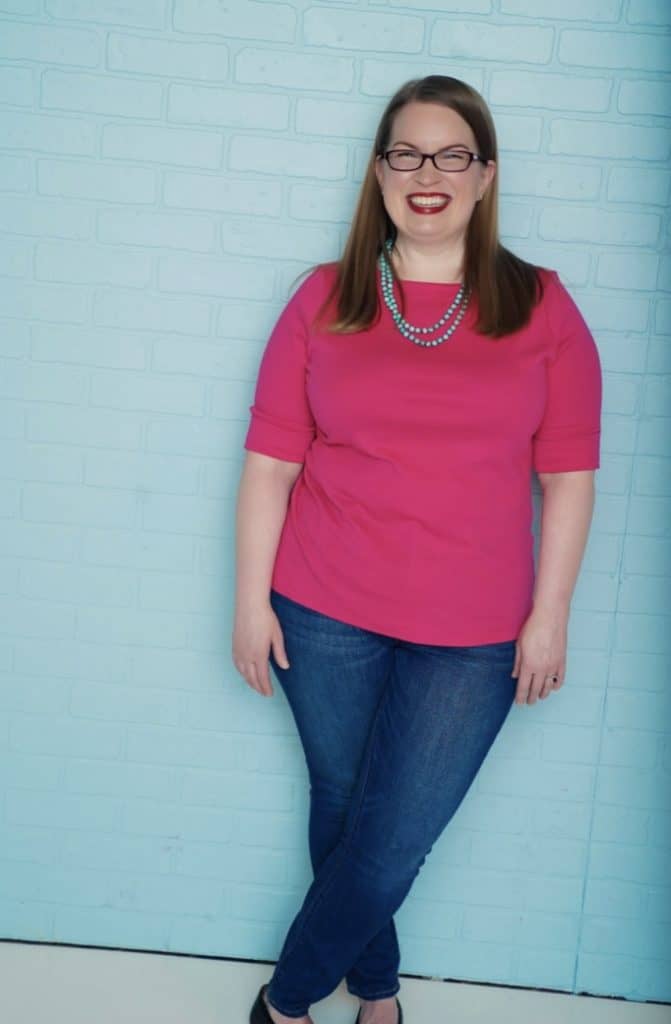 woman wearing a pink shirt, jeans, and glasses smiling in front of a blue brick wall