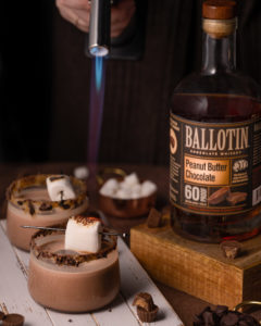torch roasting a marshmallow on top of a brown alcoholic drink next to a bottle of whiskey