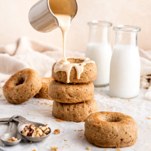 topping getting drizzled on a stack of three banana baked donuts next to two glasses of milk and a spoon of walnuts