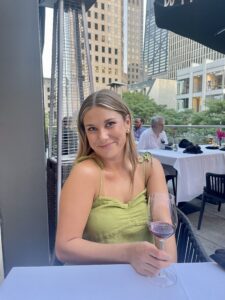 blonde woman wearing a green tank top holding a wine glass sitting at a table outdoors