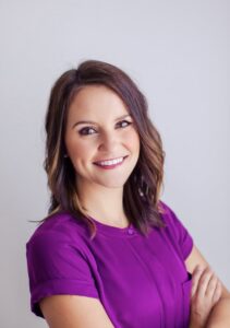 woman with brown short hair wearing a purple shirt smiling to the camera with arms crossed
