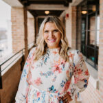 A smiling woman with long, wavy blonde hair stands in a brick corridor, wearing a floral dress with a high neckline and belted waist. The background features large windows and an inviting urban setting.