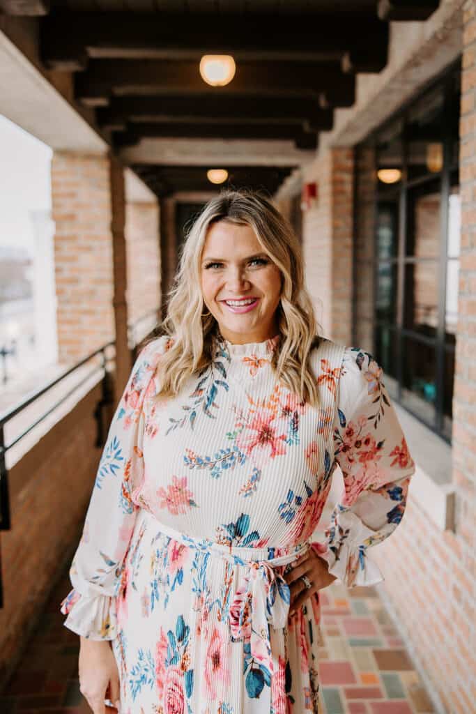 A smiling woman with long, wavy blonde hair stands in a brick corridor, wearing a floral dress with a high neckline and belted waist. The background features large windows and an inviting urban setting.