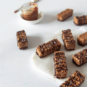 A selection of homemade chocolate cereal bars with a drizzle of chocolate on top, artistically arranged on a white plate with a jar of chocolate spread and a knife in the background, suggesting a healthy, homemade treat.