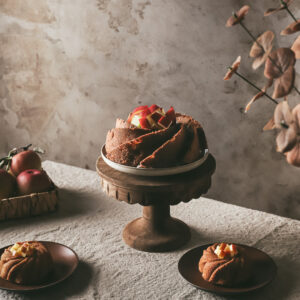 An apple bundt cake on a rustic wooden cake stand, topped with fresh apple slices arranged in a circular pattern. The cake and stand are placed on a table with a backdrop of warm, textured gray tones and a branch with autumn leaves in the background. A basket of apples is partially visible to the left.