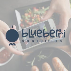 A blurred background with the words "blueberi consulting" overlaid on a semi-transparent tag. The foreground shows a hand holding a smartphone displaying the same text, with the company's logo of a stylized berry in black with a green leaf.