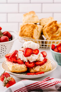A plate with strawberry shortcake, featuring a fluffy biscuit sliced in half and filled with whipped cream and sliced fresh strawberries, with more whipped cream on top. In the background, there's a colander with strawberries and a basket with additional biscuits.