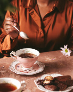 A person in an orange shirt is steeping tea, with a vintage-style pink teacup filled with tea on a saucer and a small plate of assorted chocolate biscuits beside it. A single drop of tea is dripping from the spoon they are holding, showcasing a moment of tranquility and the enjoyment of tea time.