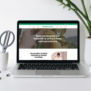 A laptop on a white desk displays a website homepage for "creative zest," which offers branding services for food entrepreneurs. The website tagline says, "Tasteful brands for humble & driven food entrepreneurs," and the site features a welcoming design with a clean, modern aesthetic. Beside the laptop are a pair of scissors and a potted plant, contributing to the creative, work-focused environment.