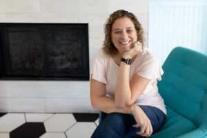 Woman with curly, shoulder-length, dirty blonde hair smiles to the camera on a teal armchair.