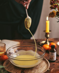 A bowl of cake batter is being mixed, with the viscous yellow mixture flowing from a spoon, illustrating the process of baking. The warm ambiance is set by a candle and flowers in the background, and ingredients like oranges and vanilla extract suggest the flavors involved in the recipe.