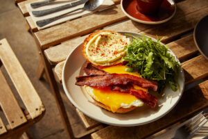 white plate on wooden table with an open-faced breakfast sandwich containing egg, cheese, bacon, and arugula