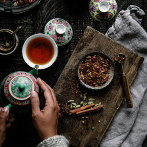 An overhead shot of a woman's hands pouring tea from a patterened kettle. There is a bowl with loose leaf tea nearby.