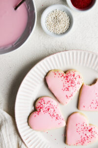 A white paper plate on a white countertop with heart-shaped sugar cookies on top. Icing and sprinkles can be seen in dishes surrounding the plate.