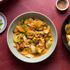 A bowl on a purple/maroon countertop. Inside is a dish with a peanut sauce, water chestnut, corn, green onion, and cabbage.