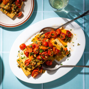 Tofu slices on a white plate with toppings of cherry tomato, capers, and basil. Plate is on a light blue countertop with cutlery nearby.