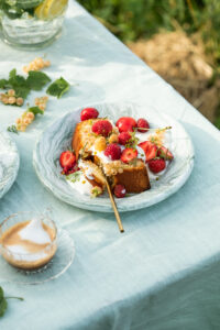 A slice of cake topped with fresh berries and a dollop of cream on a ceramic plate, set on a light blue tablecloth outdoors. A glass pitcher with lemon water and fresh mint leaves is visible in the background, creating a refreshing summer dessert setting.