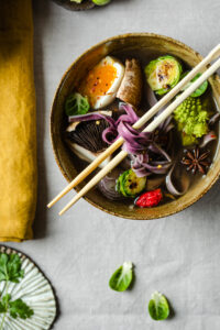 A rustic bowl filled with a vibrant, colorful ramen noodle soup featuring purple noodles, a soft-boiled egg, mushrooms, and assorted vegetables like brussels sprouts and romanesco, garnished with red chili and served with chopsticks.