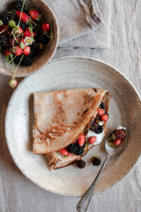 An elegant plate displaying a folded crepe filled with fresh berries and cream, accompanied by a small bowl of mixed berries. The setup is on a linen-covered table, evoking a refined, rustic brunch atmosphere.