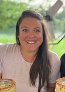 A portrait of a woman smiling warmly, outdoors with a background of greenery, holding two margaritas with salted rims.