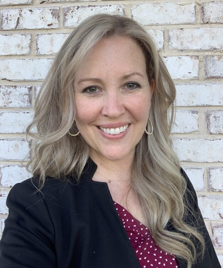 A woman with long, wavy blonde hair smiling in front of a light brick wall. She is wearing a black blazer over a red blouse with white polka dots, and gold hoop earrings. The setting is bright and casual, highlighting her friendly and approachable demeanor.