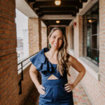 A woman with long, wavy hair stands confidently in a brick hallway. She is wearing a stylish navy blue jumpsuit with a one-shoulder ruffle detail and a cutout at the waist. She smiles warmly at the camera, with her hands on her hips.
