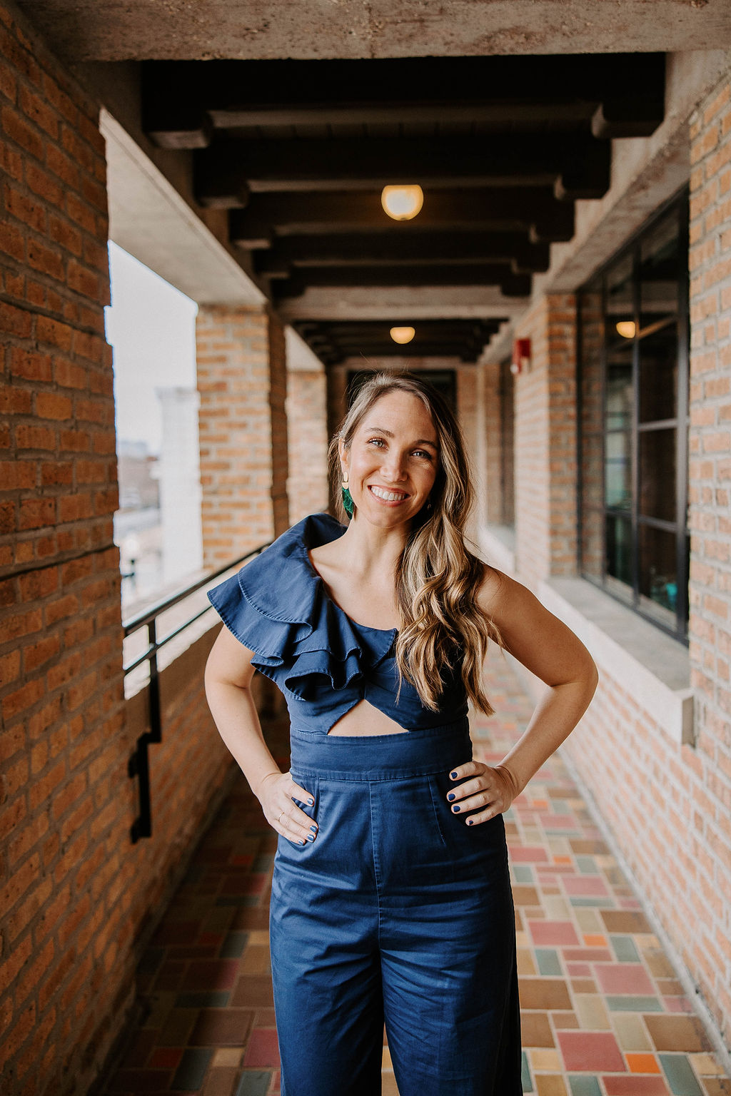 A woman with long, wavy hair stands confidently in a brick hallway. She is wearing a stylish navy blue jumpsuit with a one-shoulder ruffle detail and a cutout at the waist. She smiles warmly at the camera, with her hands on her hips.