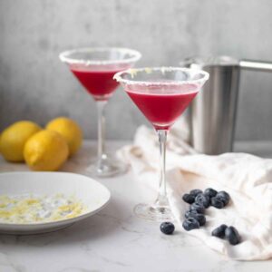 Two elegant glasses of blueberry lemon cocktails, served on a marble countertop. The drinks are vibrant red, garnished with lemon zest on the rim, and accompanied by a bowl of blueberries and lemon wedges.