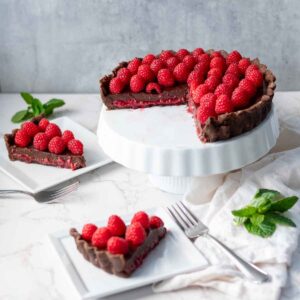 A beautifully crafted chocolate raspberry tart placed on a white pedestal cake stand. The tart is decorated with a dense layer of fresh raspberries on top, with a slice partly cut and served on a white plate with a mint garnish beside it.