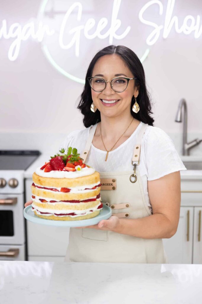 A woman with long dark hair and glasses stands in a modern kitchen, smiling while holding a beautifully layered strawberry shortcake. She is wearing a light-colored apron with her name tag, "SUGAR GEEK," and a white blouse underneath. Behind her, a neon sign reads "Sugar Geek Show."