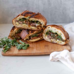 A rustic and hearty steak sandwich, sliced in half and stacked on a wooden cutting board. The sandwich is filled with tender steak slices, greens, and chimichurri sauce, displaying a savory and juicy filling.