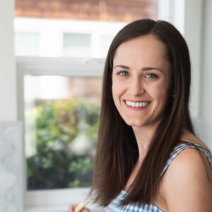 A smiling woman with long brown hair, wearing a blue and white checkered dress, standing in a bright kitchen. She looks cheerful and is holding a cooking utensil, with a pleasant outdoor view visible through the window behind her.