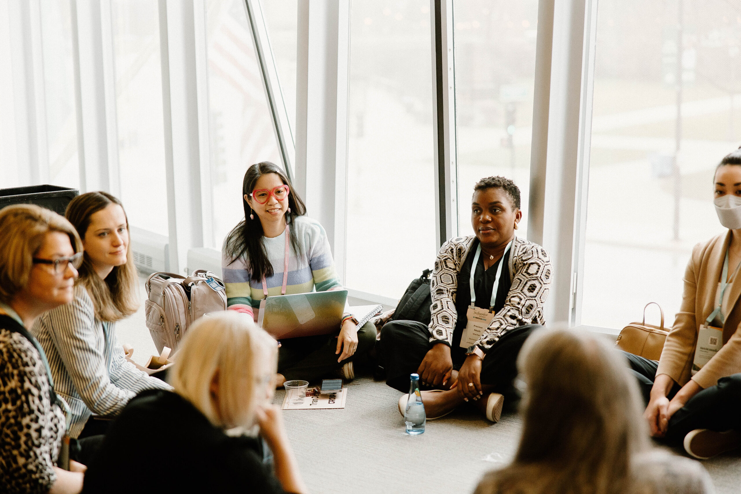 A diverse group of women is seated in a circle on the floor in a bright, spacious room with large windows. They are engaged in a discussion, some with laptops and notebooks.