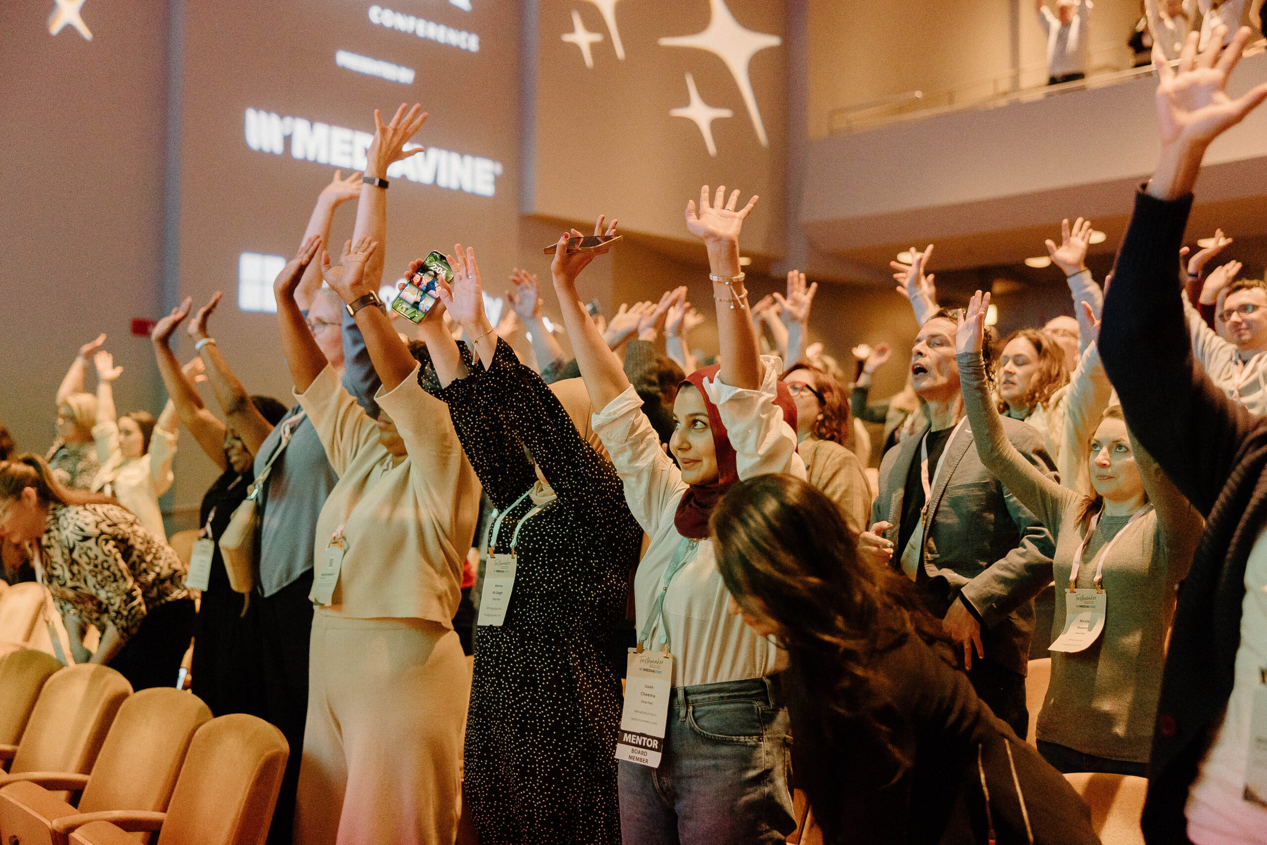 A diverse group of people, both men and women, are standing in an auditorium with their hands raised enthusiastically. The room is well-lit with a warm ambiance, and the background features logos and branding for the event, indicating it is a conference or seminar. Some attendees are wearing name badges and lanyards, and there is a palpable sense of excitement and engagement among the participants.