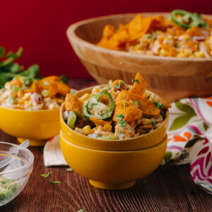 A vibrant bowl of corn salad topped with sliced jalapenos and crispy Fritos chips, served in a bright yellow bowl with a wooden bowl filled with more salad in the background.