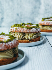 Close-up view of a gourmet cake with light green cream in between layers of cake, with a sugared top, presented on a simple white plate against a textured grey background.