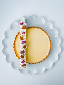 Elegant dessert presentation of a tart with a white cream topping decorated with almonds and edible purple flowers, placed on a wavy-edged blue plate.